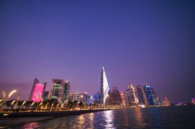 Gece körfezin üzerine çökerken uzun binalar mor gökyüzüne karşı parıldıyor..