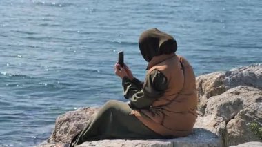 Şahıs su kenarındaki kayalıklarda oturuyor, elinde akıllı telefonla..