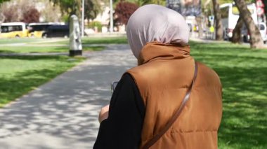 Başörtüsü takan bir kadın yeşilliklerle dolu bir parkta geziniyor..