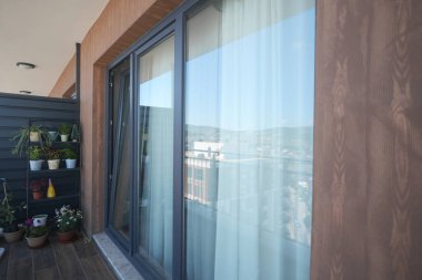 Parlak balkonda büyük pencereler, perdeler ve saksı bitkileri yer alıyor..