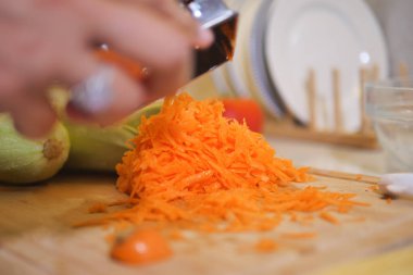 Bir insan, yanında sebzeler olan bir kesme tahtasının üzerinde parlak turuncu havuçları rendeliyor..