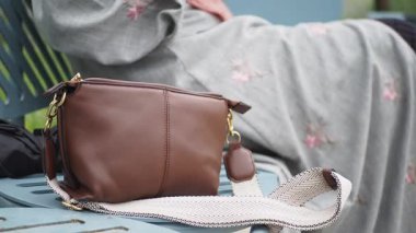 Kahverengi bir çanta bankın üzerine yerleştirilirken, bir kişi parkın yakınında dinlenir..