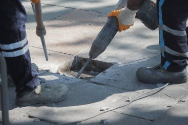 İki işçi caddedeki hasarlı kaldırımı tamir etmek için bir matkap kullanıyor..
