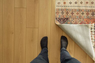 Kişi ahşap zeminde kısmen sarılmış bir halının yanında durur..