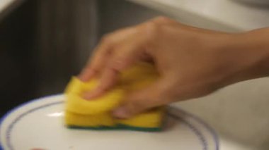Bir insan mutfakta sarı bir süngerle tabak fırçalar..