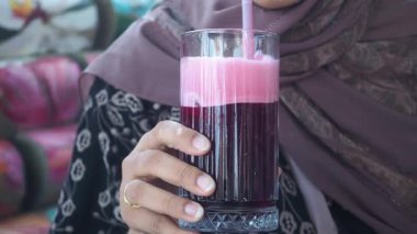 Elinde pembe köpüklü bir bardak kırmızı meyve suyu ve pipet tutan kişi..