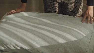 Bir insan gri bir yastığı incelerken güneş ışığı evin içine gölge düşürür..