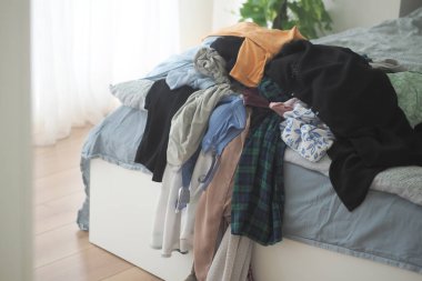 Düzensiz bir kıyafet koleksiyonu parlak bir odadaki bir yatakta duruyor..