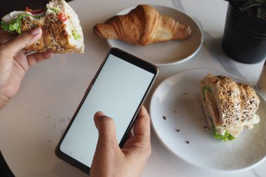 Kişi, kafede telefon kullanırken sandviç ve kruvasan yemekten hoşlanır..