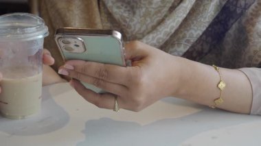 Bir kadın masada oturmuş, buzlu içkisini yudumluyor ve akıllı telefonuna odaklanıyor..