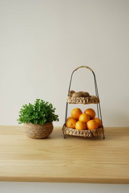 Sergide bir bitkinin yanındaki dokuma sepetlerde portakallar ve patatesler yer alıyor.