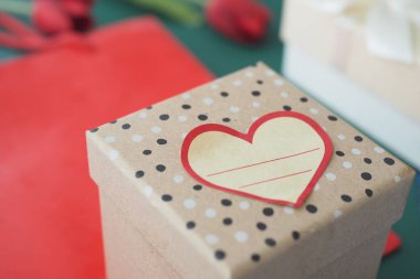 Üzerinde kalp etiketi olan bir hediye kutusu yeşil bir yüzeyde kırmızı güllerin arasında duruyor..