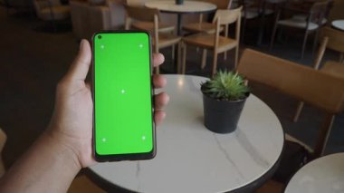 Bir kişi kafeterya ortamında yeşil ekranlı bir akıllı telefon görüntüler.