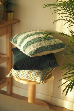 Farklı şekillerde yumuşak yastıklar, güneş ışığında parlayan ahşap bir taburede dinlenir..