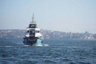 Bir feribot sakin suları aşıp mavi gökyüzünün altındaki şehir siluetine doğru yelken açar..
