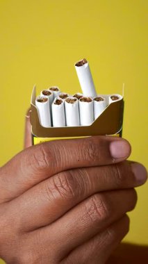 Bir el, parlak sarı bir arka planda bir paket sigara tutuyor..