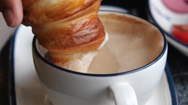 A close-up of a hand dipping a croissant into a warm cup of coffee.