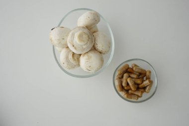 Fresh mushrooms and herbal capsules arranged in two bowls on white surface.