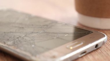Çatlak bir akıllı telefon, kahve fincanının yanındaki ahşap bir masada duruyor..