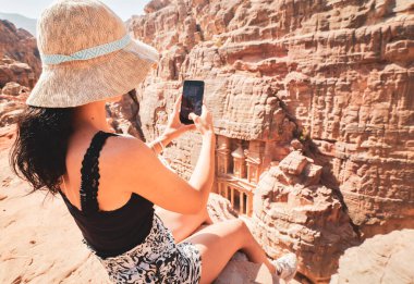 Güzel beyaz kadın turist, Petra antik kentinde Hazine ya da El-Khazneh 'in üzerinde oturarak akıllı telefon fotoğrafı çekiyor. Jordan, yedi harikadan biri. UNESCO Dünya Mirası.