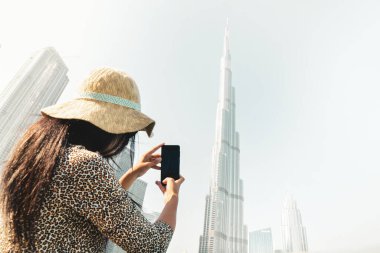 Kafkasyalı genç turist Burj Halife 'nin güneşli bir gökyüzü gününde akıllı telefonuyla mobil fotoğrafını çekiyor.