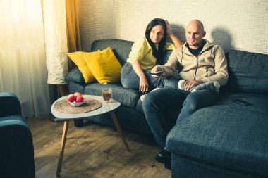 Uzaktan kumandalı beyaz bir adam kız arkadaşını kucaklıyor ve evdeki kanepede televizyon izliyor. Kanepede otururken evde televizyon izleyen sevimli bir çift.