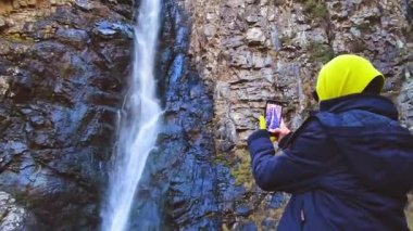 Kadın, arka planda Gveleti şelalesinin fotoğrafını çekmek için akıllı telefonu tutar ve ekrana basar