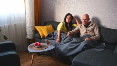 Uzaktan kumandalı beyaz bir adam kız arkadaşını kucaklıyor ve evdeki kanepede televizyon izliyor. Kanepede otururken evde televizyon izleyen sevimli bir çift.