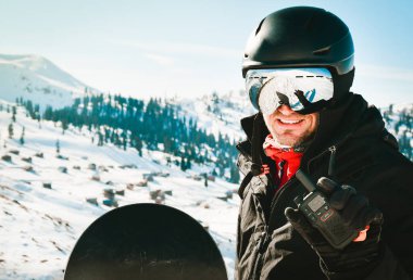 Caucasian man in black jacket and goggles with ski helmet on his head holds walkie-talkie in his hand against background of white snow