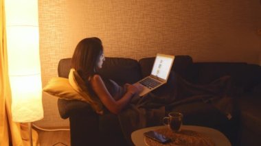 Gürcistan - 2022: Genç beyaz kadın kanepeye uzandı rahatla akıllı telefon kullan ve defter internette gezdir, yaz için hazırlan. Çevrimiçi alışveriş yaparken online fiyatları karşılaştır