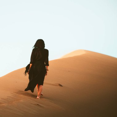 Uzun elbiseli genç bir kadın kum tepeleri boyunca yalnız başına yürüyor.