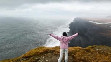 Pembe ceketli genç bir kadın dağın zirvesinde duruyor kollarını havaya kaldırıyor, mutlu ve hayattan, gençlikten ve mutluluktan sarhoş. Dyrholaey turist ve vahşi Atlantik Okyanusu