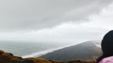Pembe ceketli genç bir kadın, uçurumun kenarına doğru düşünceli bir şekilde yürüdü. İzlanda 'da okyanus dalgalarını izledi. Güney Vik' teki Black sand plajı. Kadın sağlığı ve refahı.