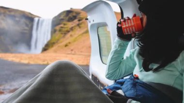 Çekici genç bir bayan, İzlanda 'da, Skogafoss şelalesinin tadını çıkarırken karavanının içinde battaniyeyle oturup kahve içiyor. Yol gezisi çevresi İzlanda doğası ziyareti