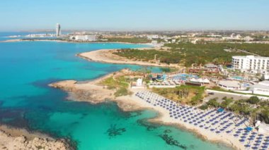 Ayia Napa, Kıbrıs Rum Kesimi - 15 Nisan 2023: Lüks otel binalarının üzerinde ada yeşili manzaralı havuz kenarlı uçuş. Kıbrıs 'ın en ünlü beyaz kumu - Nissi plajı