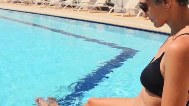 度假时 穿着黑色比基尼的后视镜美女坐在酒店度假胜地的游泳池边 游客们享受阳光 假日里阳光灿烂的日子 在游泳池边晒日光浴放松 — 图库视频影像