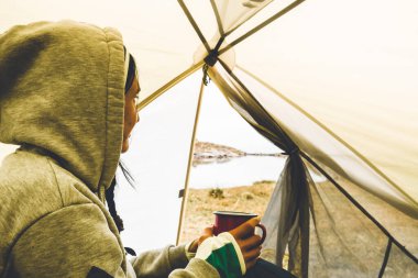 Smiley young caucasian woman hold camping mug and drink in green tent outdoors in nature with viewpoint panorama of alpine lake in autumn