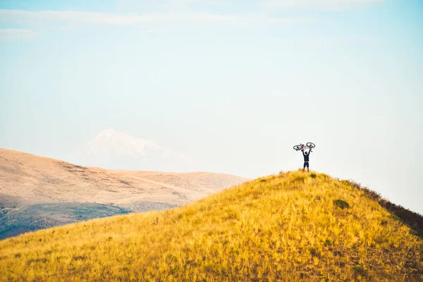Isolated fit young caucasian male person lift bicycle up on hill with mountains background outdoors in caucasus mountains. Achievement , inspiration, challenge and determination concept