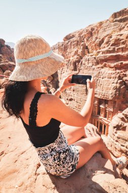 Güzel beyaz kadın turist, Petra antik kentinde Hazine ya da El-Khazneh 'in üzerinde oturarak akıllı telefon fotoğrafı çekiyor. Jordan, yedi harikadan biri. UNESCO Dünya Mirası.