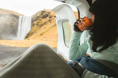 Turist kadın oturup kahve içiyor. Kiralık karavanın içindeki açık kapılı doğa manzarasının tadını çıkarıyor. Skogafoss Şelalesi, İzlanda. Tek başına İzlanda 'yı gezmek. Resmi kamp alanı olarak kalın.
