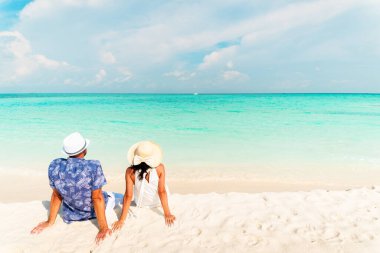 Şapkalı güzel bir turist çift, tropikal bir plajda oturur ve yaz tatillerinde turkuaz okyanusun tadını çıkarırlar.