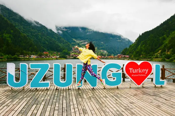 stock image Young caucasian woman tourist jump out of joy on travel funny pose outdoors by Uzungol billboard sign in Turkey. Travel joy and freedom destination concept
