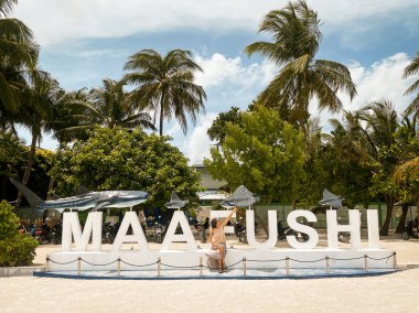 Maafushi, Maldivler - 7 Mayıs 2024: Turist kadın ünlü Maafushi 'nin yanında oturuyor tropik tatil adasında hoş geldin işareti