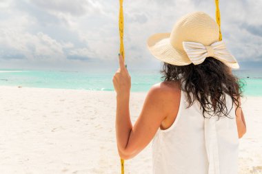 Hasır şapkalı genç bir kadın tropikal okyanus kıyısında salıncakta sallanıyor ve boş fotokopi alanı arıyor. Kız bakış açısı. Tatil konsepti. Turkuaz suyla huzur manzarası