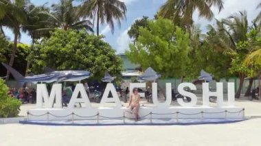 Maafushi, Maldivler - 7 Mayıs 2024: Turist kadın ünlü Maafushi 'nin yanında oturuyor tropik tatil adasında hoş geldin işareti