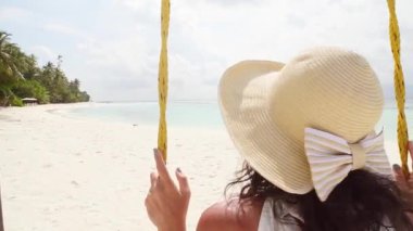 Hasır şapkalı genç bir kadın tropik okyanus kıyısında salıncakta sallanıyor ve boş fotokopi alanına bakıyor. Beyaz giysili kız. Tatil konsepti. Turkuaz suyla huzur manzarası
