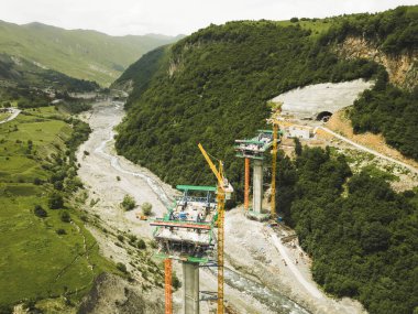 Kazbegi, Gürcistan - 7 Temmuz 2024: Çinli inşaatçılar tarafından inşa edilen yeni otoyol projesinin inşaat sahasında havadan görüş vinçleri ve köprü temel direkleri. Çığ baypas tüneli. Tbillisi - Lars