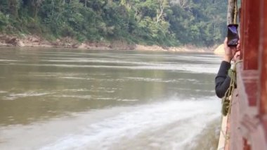 Luang Prabang, Laos - 15 Ocak 2024: Ünlü yavaş tekne yolcuları tatillerde akıllı telefon film tatili deneyimine sahipler. Laos 'tan Tayland' a Manzaralı nehirde yavaş tekne gezisi