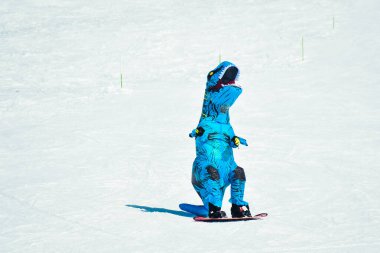 Gudauri, Gürcistan - 15 Şubat 2024: önde kayakçı mavi dinozor kostümlü snowboard giyer ve kış tatillerinde kayak merkezinde eğlenir.