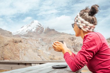 Kafkasyalı genç mutlu turist kadın dağda kafeteryada oturuyor sıcak çikolata içiyor Kazbek dağlarının zirvesini seyrediyor Kazbegi ulusal parkında. Gürcistan 'a sonbaharda seyahat
